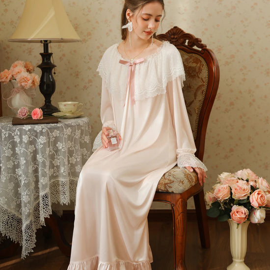 Seated front view of the light pink long nightgown showing its wide white lace collar, pink ribbon bow, and ruffle hem