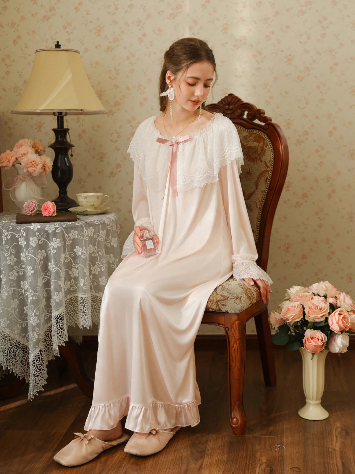 Seated front view of the light pink long nightgown showing its wide white lace collar, pink ribbon bow, and ruffle hem