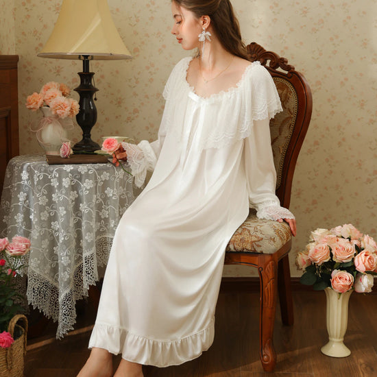 Seated three-quarter view of the white long nightgown with a broad scalloped lace collar, white ribbon bow, and ruffled hem