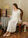 Seated three-quarter view of the white long nightgown with a broad scalloped lace collar, white ribbon bow, and ruffled hem