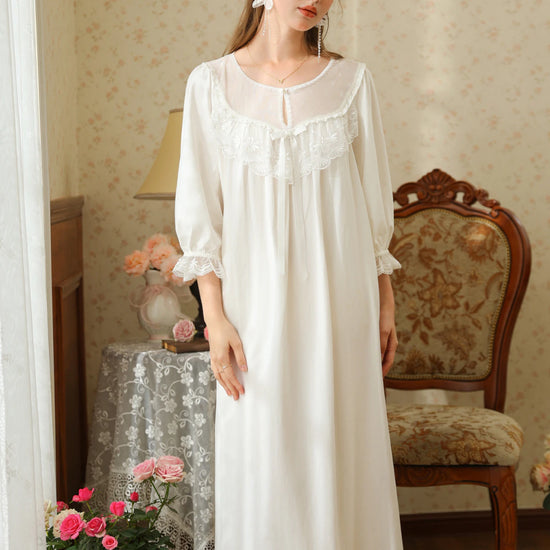 Front view of the white cotton nightgown showing its sheer embroidered yoke, scalloped lace bib, satin bow, and midi hem