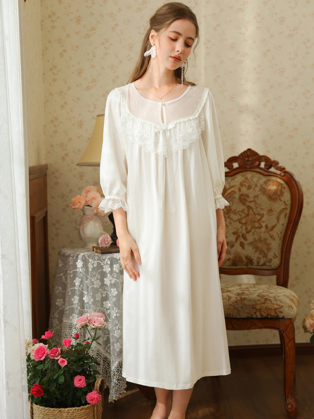 Front view of the white cotton nightgown showing its sheer embroidered yoke, scalloped lace bib, satin bow, and midi hem