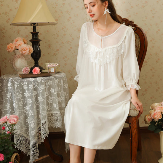 Seated view of the white cotton lace yoke nightgown showing its loose skirt drape and long satin ribbon bow
